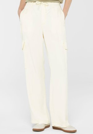 Person trägt lockere, cremefarbene Cargo-Hosen mit Kordelzug in der Taille und Seitentaschen, kombiniert mit weißen Turnschuhen und einem in die Hose gesteckten Oberteil.