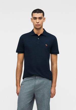 Bărbat tânăr purtând un tricou polo bleu închis cu un mic logo brodat cu elan și pantaloni gri deschis, stând pe un fundal simplu.