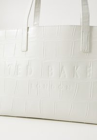 Weiße krokodilpräge Tote-Bag mit zwei Griffen, mit dem eingeprägten Markennamen "TED BAKER LONDON" auf der Vorderseite. Glatte Textur.