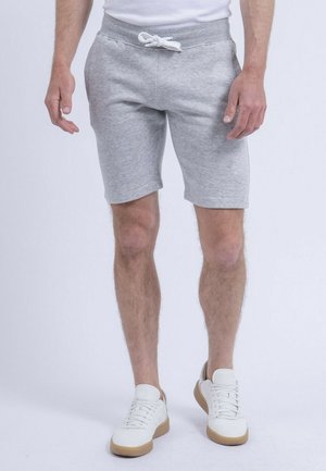 Homme portant un short gris à cordon de serrage et des baskets blanches, debout devant un fond clair uni.