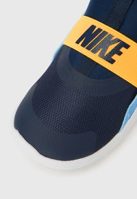 Gros plan sur une basket Nike bleu marine avec un bout texturé, des accents bleu clair et une sangle orange vif ornée du logo Nike noir.