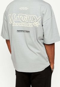 Šedé bavlněné tričko s volným střihem. Velký bílý grafický text na zadní straně, obsahující "Multiply Apparel" a další detaily značky.