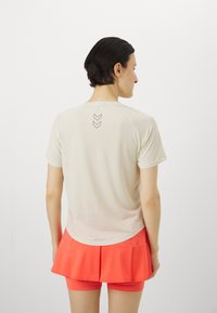 Beige sportskjorte med korte ermer og avrundet søm, med en mørk logo-design. Kombinert med en lys korallfarget skorts.