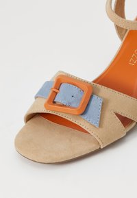 Sandalia de ante beige con tira de cuero azul y hebilla cuadrada naranja. La plantilla es naranja y tiene una textura suave.