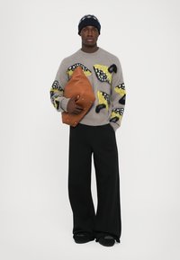 Pulover gri tricotat cu modele abstracte în galben și negru, purtat împreună cu pantaloni evazați negri. Ținând o geantă oversized de culoare maro.
