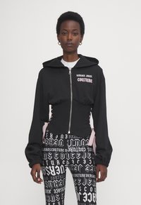 Čierna skrátená mikina s ružovými bočnými akcentmi a predným zipsom, na ktorej je vyšívané "Versace Jeans Couture" v bielej farbe. Vzorované čierne nohavice.
