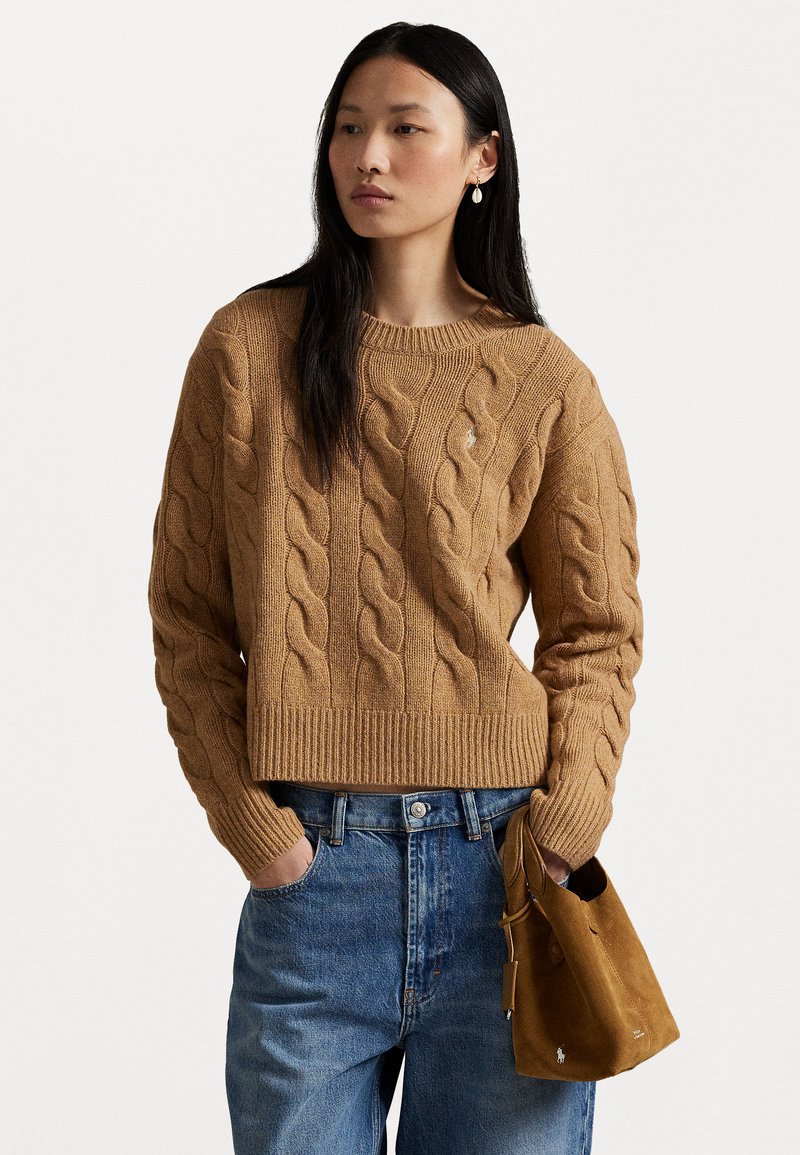 Jersey de punto acanalado en marrón claro, con dobladillo y puños acanalados. Combinado con jeans azul de cintura alta y sosteniendo un pequeño bolso de ante.