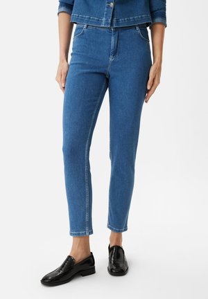 Niebieskie dżinsy o slim fit, z średnim stanem, przednimi kieszeniami i czystym wykończeniem. Noszone z czarnymi butami na slip-on.