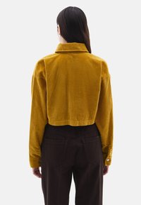 Zkrácená hořčicová kordová bunda s límečkem a dlouhými rukávy, s vertikálně pruhovanou texturou. Nosit s tmavě hnědými kalhotami.