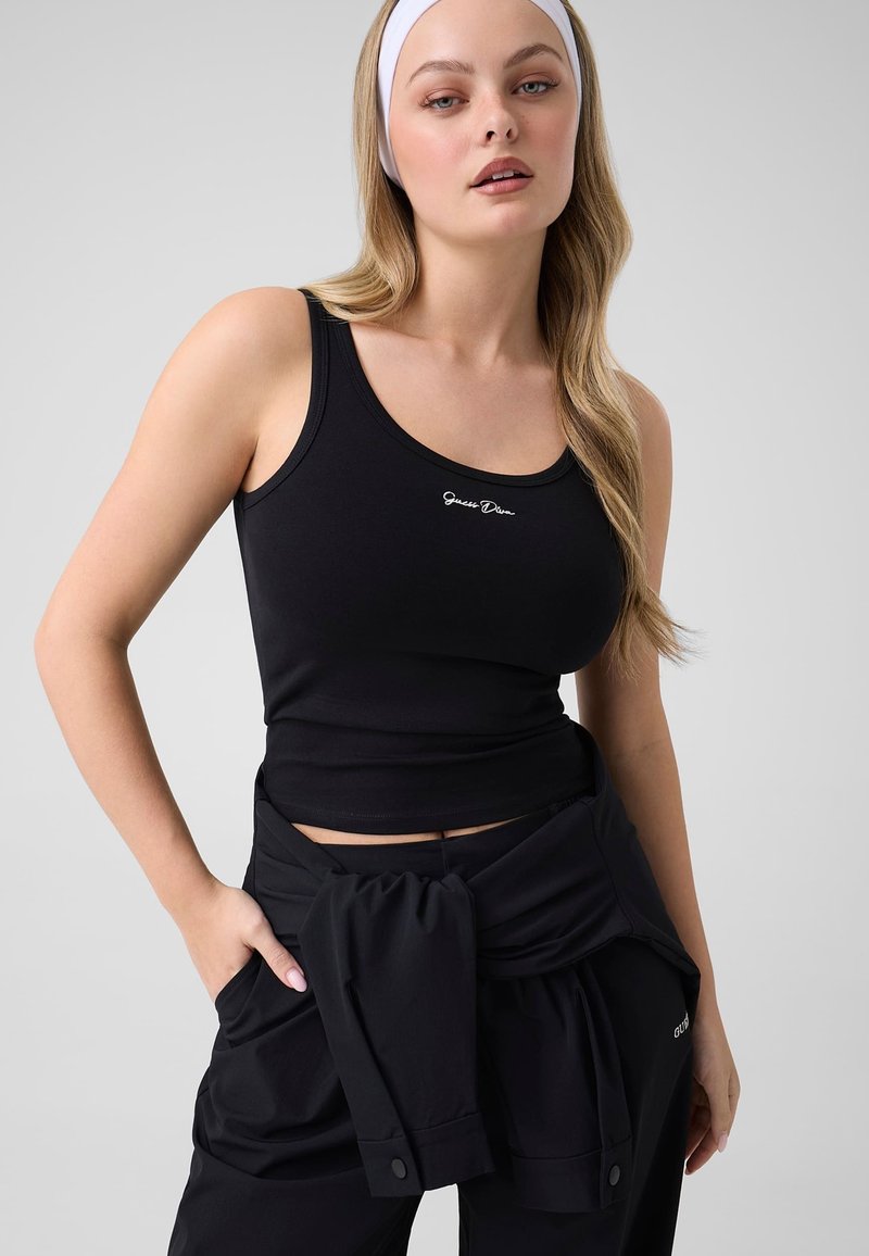 Femme aux longs cheveux blonds portant un bandeau blanc, un débardeur noir et un pantalon noir avec une veste nouée à la taille, debout devant un fond uni.
