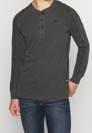 Homme portant une chemise henley à manches longues gris foncé avec trois boutons et un jean bleu foncé, debout devant un fond uni.