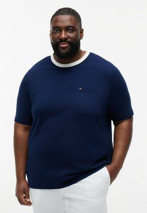 Mann mit kurzen Haaren und Bart, trägt ein marineblaues T-Shirt mit weißem Kragen und weißen Hosen, steht mit einer Hand in der Tasche.