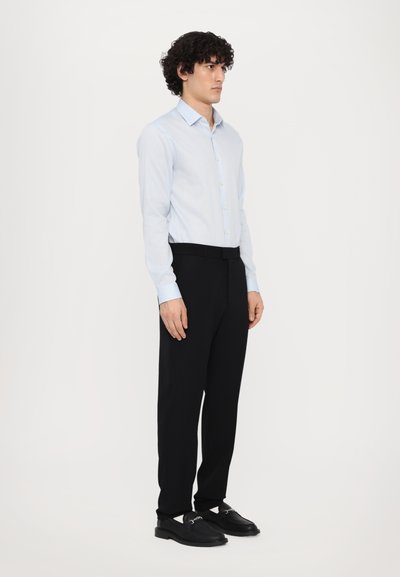 Chemise bleu clair à manches longues, col ouvert ; pantalon noir ajusté ; chaussures noires avec une boucle décorative.
