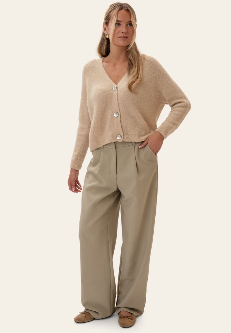 Femme portant un cardigan beige boutonné, un pantalon large beige et des chaussures plates marron, debout avec une main dans la poche.