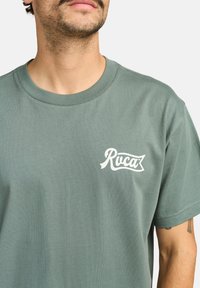 Vihreä puuvillapaita, jossa on pyöreä kaula-aukko, lyhyet hihat ja valkoinen "Ruca" -logo painettuna vasempaan rintakappaleeseen. Pehmeä rakenne.