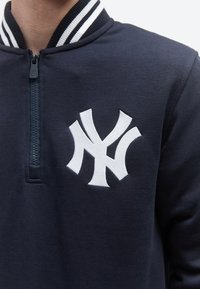 Pulover bleumarin cu fermoar de un sfert și logo NY brodat alb pe partea stângă a pieptului, purtat de o persoană.