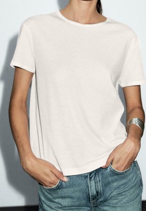 Personne portant un t-shirt blanc uni à manches courtes et un jean bleu, les mains dans les poches, avec un bracelet manchette argenté au poignet gauche.