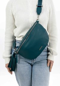 Tyrkysová kožená crossbody taška s texturovaným povrchem, oválným tvarem a stříbrnými doplňky. Obsahuje akcent s třásněmi a nastavitelný popruh.