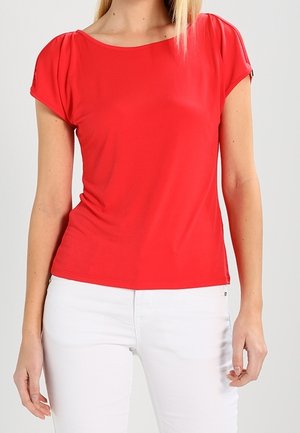 Mujer con una camiseta roja ajustada de manga corta y pantalones blancos, de pie con los brazos relajados a los lados contra un fondo liso.