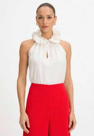 Donna che indossa un top bianco senza maniche con colletto arricciato e pantaloni rossi a vita alta, in piedi davanti a uno sfondo bianco uniforme.