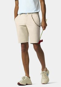 Beige shorts met een gladde voorkant, riemlussen en zijzakken. Gecombineerd met lichtgekleurde sneakers. Een rode bloem wordt vastgehouden.