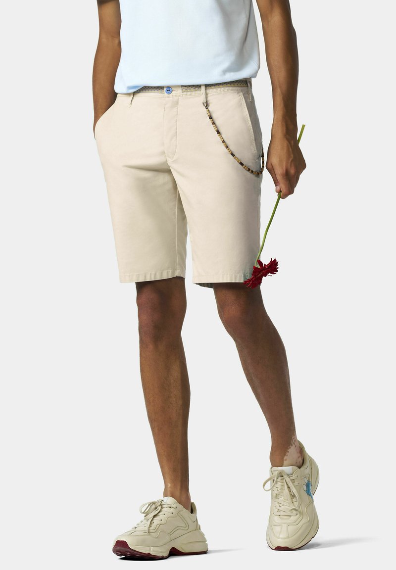 Beige shorts met een gladde voorkant, riemlussen en zijzakken. Gecombineerd met lichtgekleurde sneakers. Een rode bloem wordt vastgehouden.