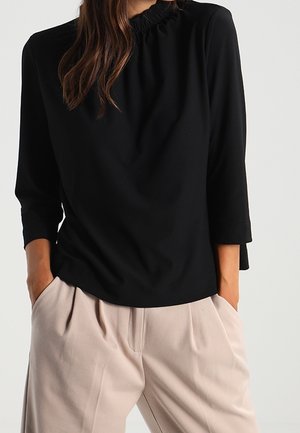 Woman wearing a black long-sleeve top with a gathered neckline and beige pleated wide-leg trousers, hands in pockets.