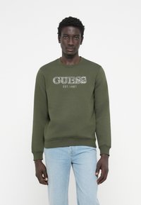Olivovo zelená mikina s logom "GUESS EST. 1981" v bielej farbe, s rebrovanými manžetami a lemom, v kombinácii so svetlo modrými džínami.