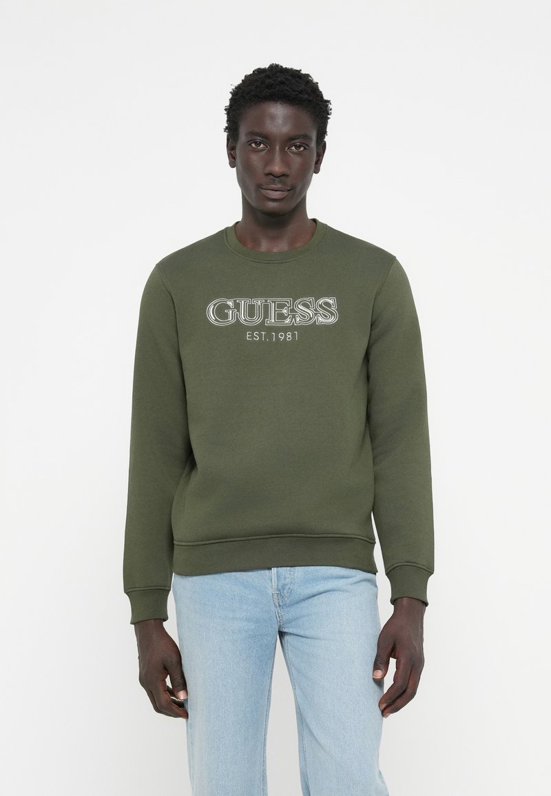 Olivovo zelená mikina s logom "GUESS EST. 1981" v bielej farbe, s rebrovanými manžetami a lemom, v kombinácii so svetlo modrými džínami.