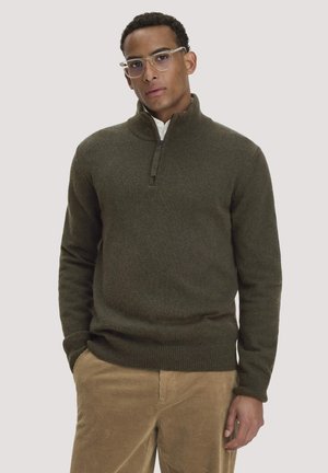 Mann mit klaren Brillen, dunkelgrünem Pullover mit Viertel-Reißverschluss und hellbraunen Cordhosen vor einem einfarbigen hellen Hintergrund.