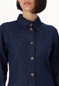 Tummansininen farkkupaita, jossa on kaulustoppi, kuusi nappia edessä, muotoillut saumat ja kapea siluetti.