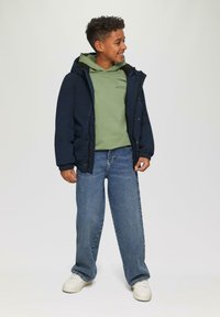 Giacca isolante blu navy con cappuccio sopra un felpa verde, abbinata a jeans larghi blu e sneakers bianche; presenta polsini elastici e tasche.