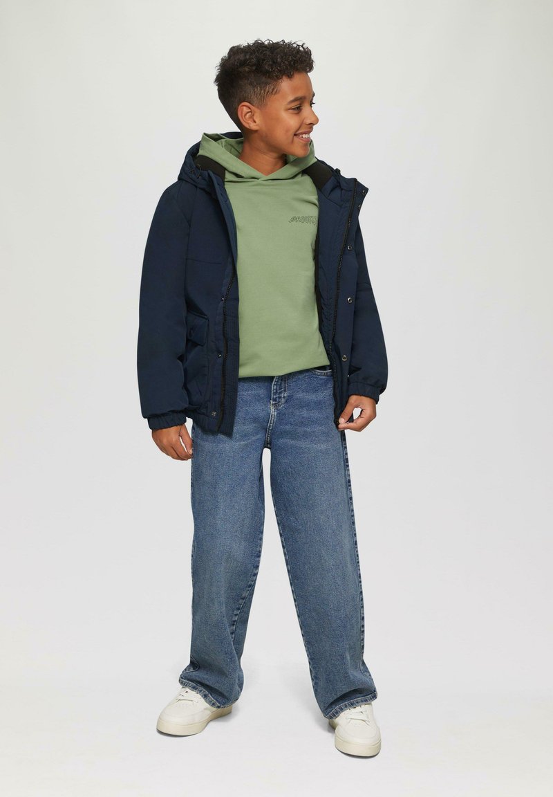 Giacca isolante blu navy con cappuccio sopra un felpa verde, abbinata a jeans larghi blu e sneakers bianche; presenta polsini elastici e tasche.