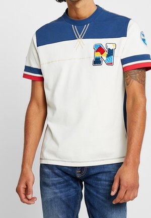 Weißes T-Shirt mit kurzen Ärmeln und blauen Schulterpartien, mit einem bunten "N"-Aufnäher sowie roten und blauen Streifenakzenten an den Ärmeln.