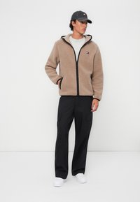 Giacca di pile beige con zip frontale, dettagli neri e cappuccio. Abbinata a pantaloni cargo neri e scarpe slip-on bianche.