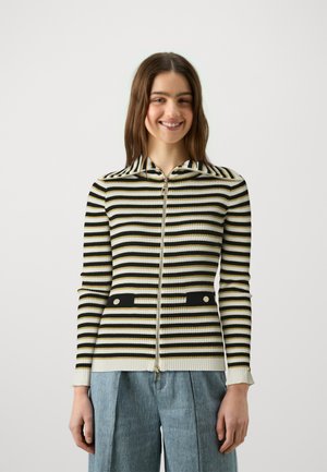 Cárdigan de cremallera a rayas blanco y negro con textura acanalada, que presenta un cuello y bolsillos negros contrastantes con detalles dorados.