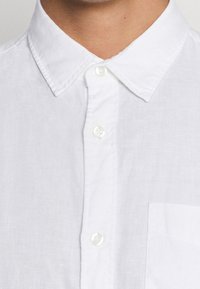 Camicia bianca in lino con collo e chiusura a bottoni, dotata di taschino sul petto e di un tessuto strutturato. Design pulito e semplice.