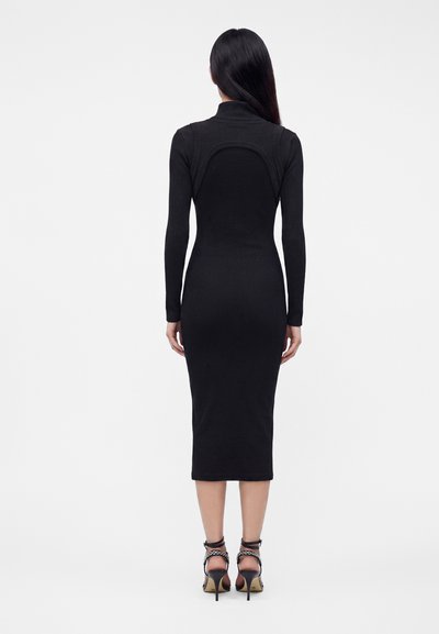 Femme aux longs cheveux noirs portant une robe midi noire ajustée à manches longues et des sandales noires à talons hauts, debout de dos.