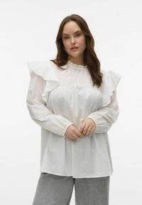 Blouse blanche avec des manches à volants, corsage plissé et texture à pois. Tissu doux, col montant et poignets froncés.