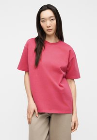 Νεαρή γυναίκα με μακριά σκούρα μαλλιά, ντυμένη με ροζ άνετο μπλουζάκι με κοντά μανίκια και μπεζ παντελόνι, στέκεται μπροστά σε απλό φόντο.