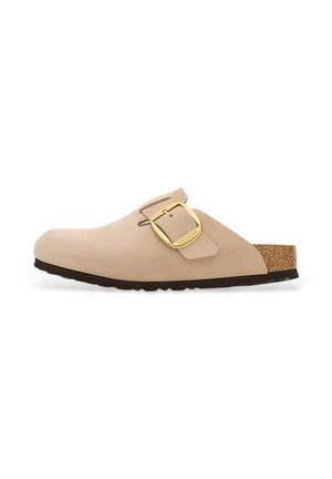 Zueco beige con forma redondeada, hecho de un material suave. Presenta una gran hebilla dorada y una plantilla de corcho, con una suela de goma negra.