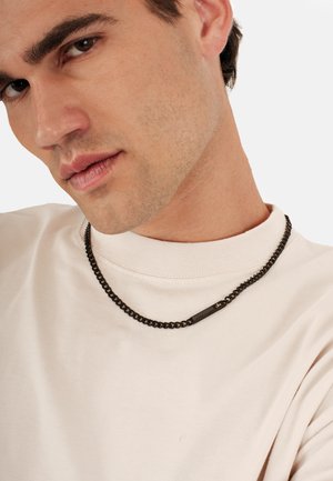 Svart kedjehalsband med platta länkar, prytt med en rektangulär detalj med guldaccent. Bärs över en ljusbeige sweatshirt.