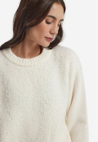 Överdimensionerad, krämfärgad stickad tröja med texturerad kropp och ribbad rund halsringning. Funktioner inkluderar sänkta axlar och vida ärmar. Mjuk och mysig kvalitet.