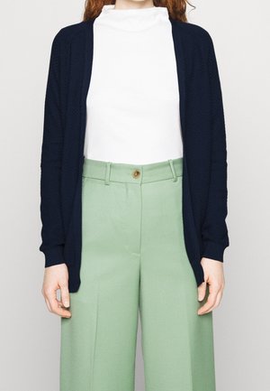 Persoon die een wit topje met hoge hals, een marineblauwe cardigan met open voorkant en lichtgroene, wijde, hoog taillebroeken met een bruine knoop draagt.