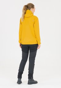 Gul vandtæt jakke med en tætsiddende hætte, parret med sorte bukser med knælapper og sorte gummistøvler. Enkel, funktionel design.