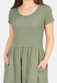 Olivengrünes Kleid mit einem gerippten Oberteil, einem Rundhalsausschnitt und kurzen Ärmeln, einem schwingenden Rock mit Taschen und einer zerknitterten Textur.