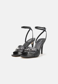 Black high-heeled sandals with a patent leather finish, open toe, ankle strap, and a metallic chain accent across the front.