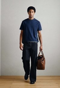 Tumsā zila īss piedurkņu krekls, brīva stila tumši džinsa bikses, melni apavi un brūna ādas soma ar rokturiem. Vienkāršs, minimālistisks dizains.