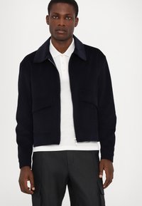 Navy corduroy jacket with a zip front, pointed collar, and two front pockets. Layered over a white polo shirt, sharp cut and texture visible.