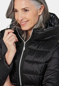 Schwarze Pufferjacke mit hohem Kragen und Reißverschluss, ausgestattet mit gestepptem Design und glänzendem Stoff. Silberne Hardware-Akzente.